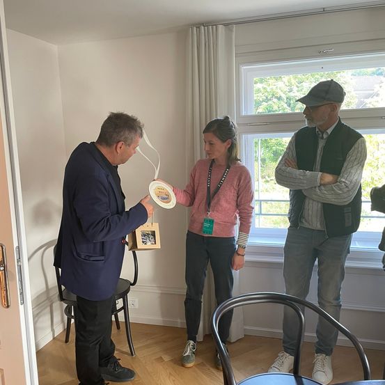 Ein Mann in einem blauen Anzug hält eine dekorative Platte, während eine Frau in einem rosa Pullover und ein Mann in einem Weste hinter ihm in einem Raum mit Holzböden und einem Fenster stehen, das Bäume zeigt.