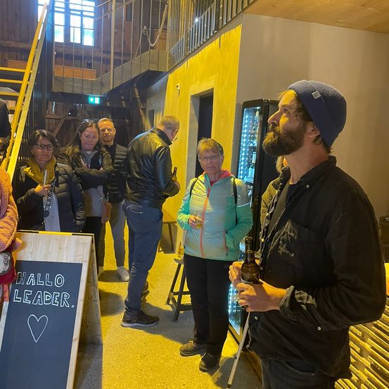 Eine Gruppe von Menschen steht in einem Raum mit gelber Wand, einige halten Getränke. Ein bärtiger Mann hält eine Flasche und eine Dose, während ein anderer Mann mit Mütze ihn ansieht. Ein Schild steht 'HALLO LEADER'.