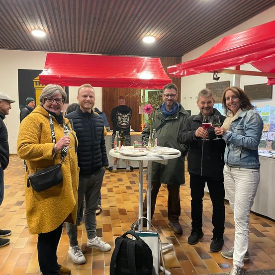 Fünf Erwachsene stehen lächelnd bei einer Indoor-Veranstaltung zusammen. Sie halten Weingläser. Ein Tisch mit einer Blume und Tellern steht in der Mitte. Rote Zelte sind über ihnen.