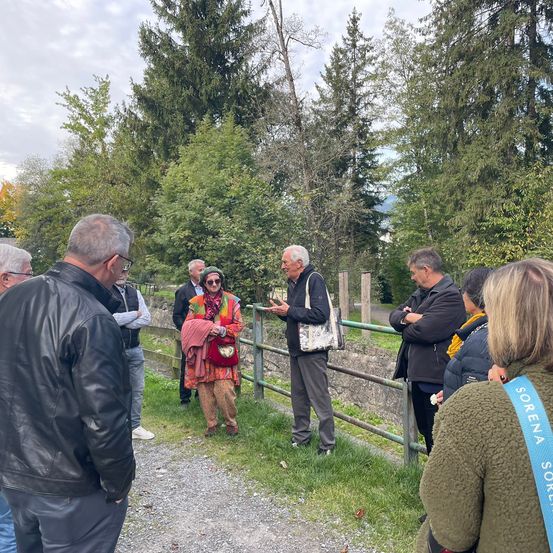 Eine Gruppe von Menschen, einige mit Brille, steht in einem Park mit Bäumen und einem Zaun. Eine Frau in einem roten Kleid hält eine rote Tasche und spricht mit einem älteren Mann.