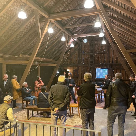In einer Scheune versammeln sich mehrere Personen. Ein Mann steht auf einem Tisch, während andere auf Bänken sitzen. Ein Schreibtisch und Stühle stehen in der Nähe. Holzbalken und Lampen schmücken die Decke.