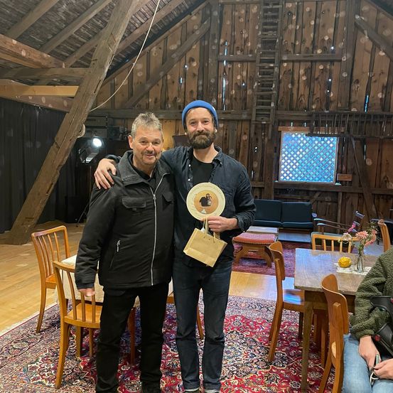 Zwei Männer stehen in einem rustikalen Raum mit Holzbalken. Einer hält einen Pokal, und sie lächeln beide. Ein Tisch, Stühle und eine Leiter sind im Hintergrund.