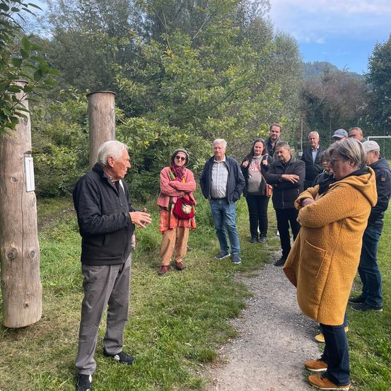 Eine Gruppe von Menschen steht in einem Park, wobei ein Mann spricht und die anderen aufmerksam zuhören. Sie sind von Bäumen und Gras umgeben.