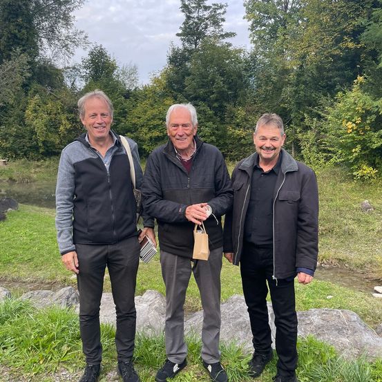 Drei Männer stehen in einem grasbewachsenen Bereich, lächeln und posieren für ein Foto. Der Mann in der Mitte hält einen kleinen Papierbeutel. Sie tragen alle schwarze Jacken und Hosen. Der Hintergrund hat einen kleinen Fluss und Bäume.