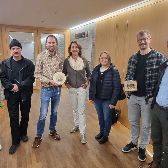 Eine Gruppe von sechs Personen steht in einem Raum mit Holzböden und Schränken. Der Mann in der Mitte hält eine Plakette und alle lächeln.