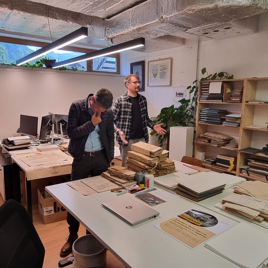 Zwei Männer sind in einem Büroraum mit einem Tisch voller Dokumente und einem Laptop. Ein Mann hat seine Hand im Gesicht. Im Hintergrund befinden sich Bücherregale und eine Pflanze.