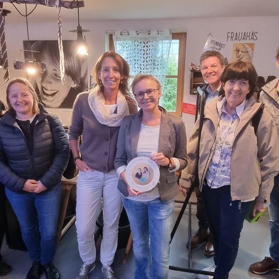 Eine Gruppe lächelnder Erwachsener steht in einem Raum. Eine Frau hält ein gerahmtes Bild, während andere zuschauen. Hinter ihnen ist ein Bild eines lächelnden Mädchens und ein Fenster mit Vorhängen zu sehen.