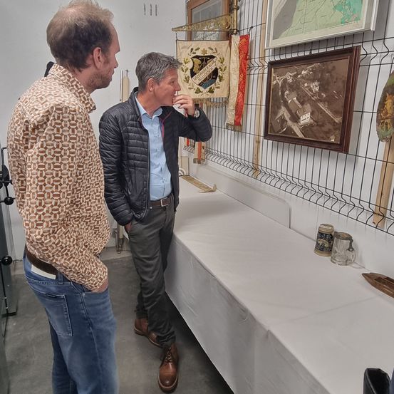 Zwei Männer sind in einem Raum mit einem langen weißen Tisch, einer trägt ein gemustertes Hemd und der andere eine Jacke. Sie scheinen zu reden. Es gibt Bilder und Gegenstände an der Wand.