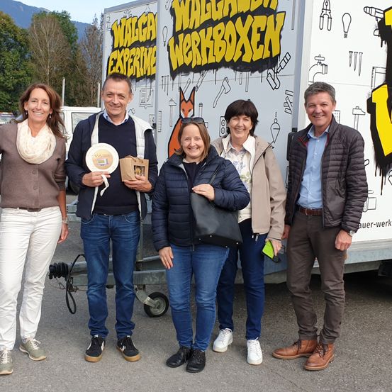 Fünf Personen stehen vor einem großen Fahrzeug mit 'WALGAU WORKBOXEN' darauf geschrieben. Der Mann auf der linken Seite hält einen Teller und einen Papierbeutel. Die Frau auf der linken Seite trägt ein Tuch. Die Frau in der Mitte trägt eine Sonnenbrille auf dem Kopf. Der Mann auf der rechten Seite trägt eine Uhr.