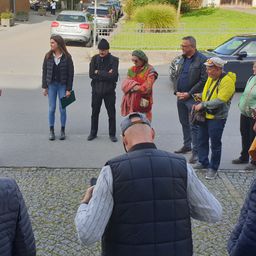 Eine Gruppe von Menschen steht auf einer Straße mit geparkten Autos und Grünpflanzen im Hintergrund. Sie scheinen eine Diskussion zu führen.