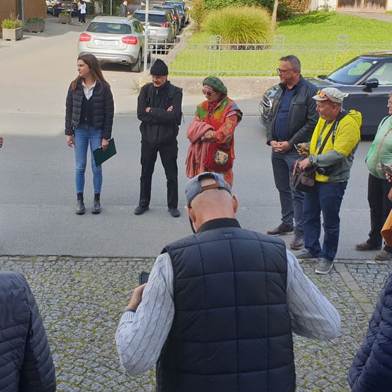 Eine Gruppe von Menschen steht auf einer Straße mit geparkten Autos und Grünpflanzen im Hintergrund. Sie scheinen eine Diskussion zu führen.