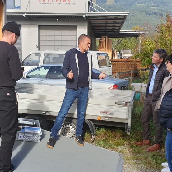 Mehrere Männer stehen vor einem Gebäude mit einem Auto und einem Lastwagen im Hintergrund. Ein Mann in einer blauen Jacke gestikuliert, während ein anderer Mann in einer schwarzen Jacke zuhört.
