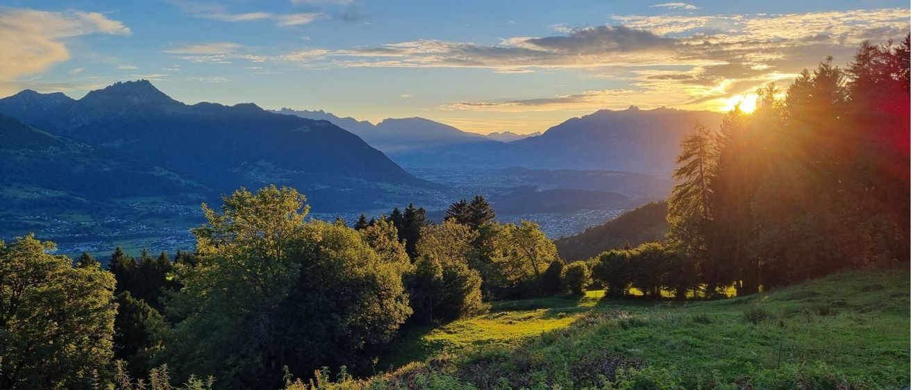 Eine ruhige Berglandschaft bei Sonnenuntergang mit Bäumen und einem Tal darunter, unter einem teilweise bewölkten Himmel.