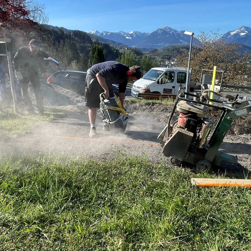 Zwei Männer schneiden mit einer Kettensäge Gras. Ein Mann beugt sich nach vorne, während der andere zusieht. In der Nähe steht ein geparkter Van und ein Baufahrzeug. Dahinter befinden sich Bäume und Berge.
