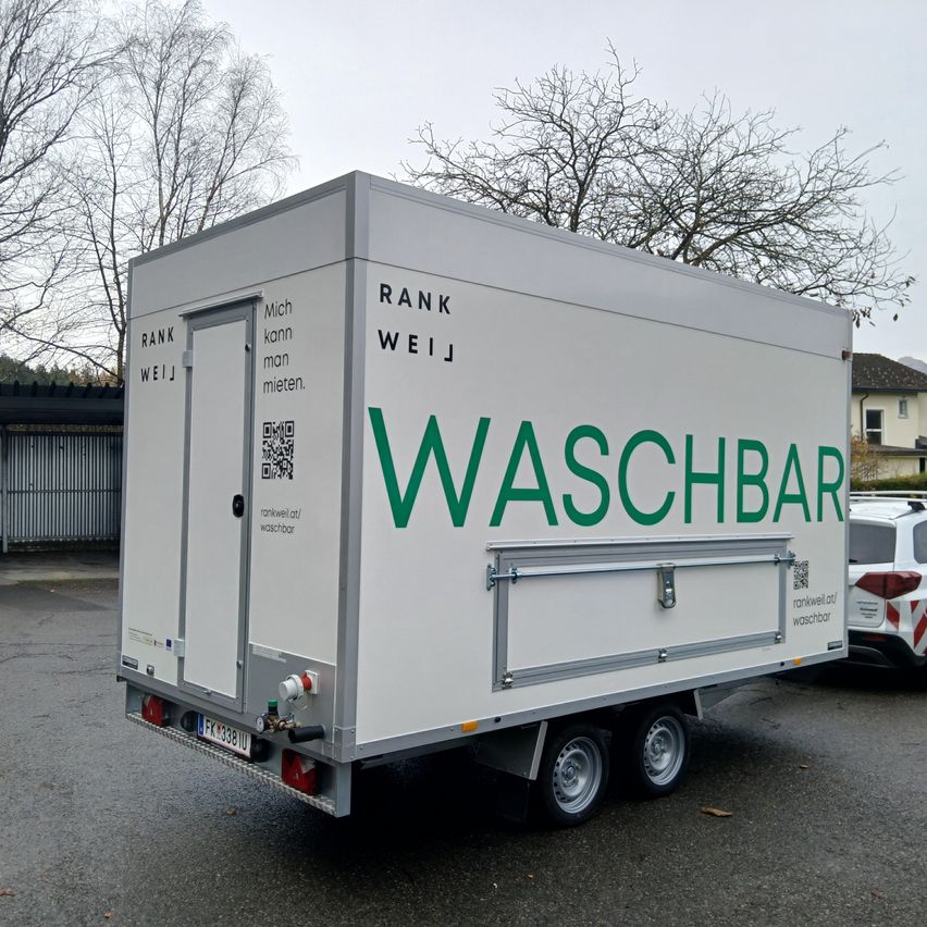 A white trailer reads 'WASHBAR' with a door and a QR code. It is parked on the road. Behind it, there is a building with trees.