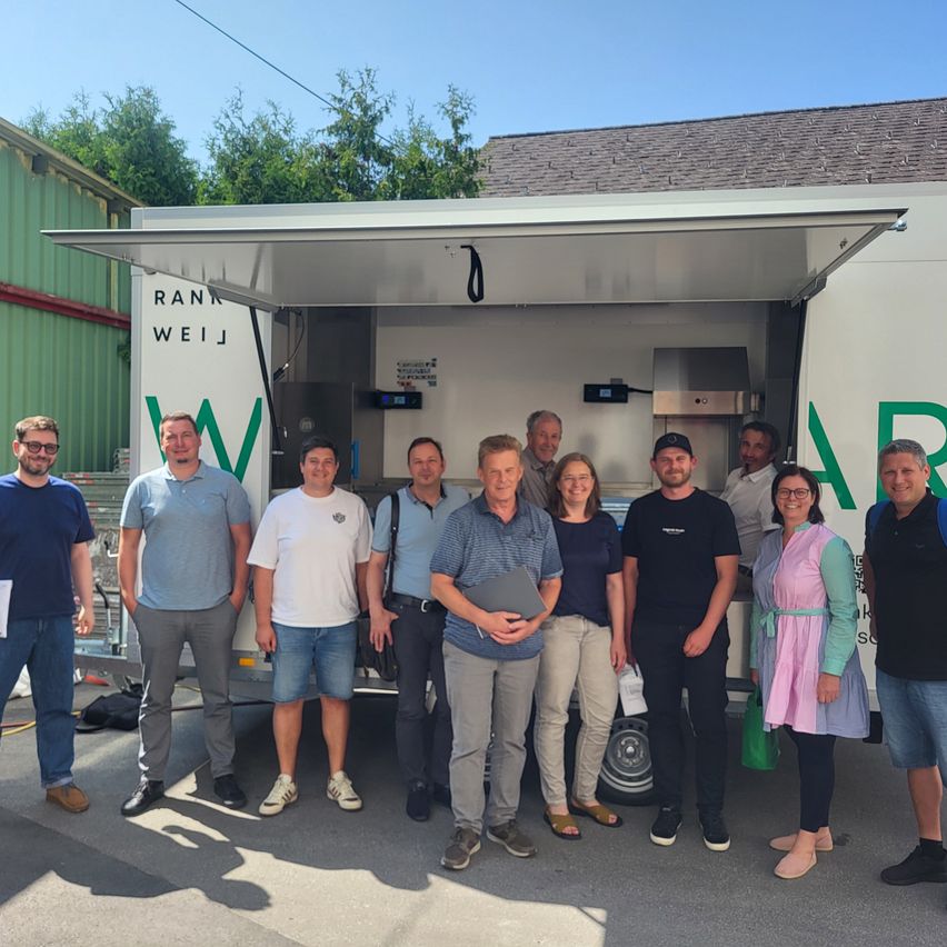 A group of individuals stands outside a white truck with the text 'RANK WEIß'. They are dressed casually, with some holding papers. Behind them, there is a house with a roof and a tree.