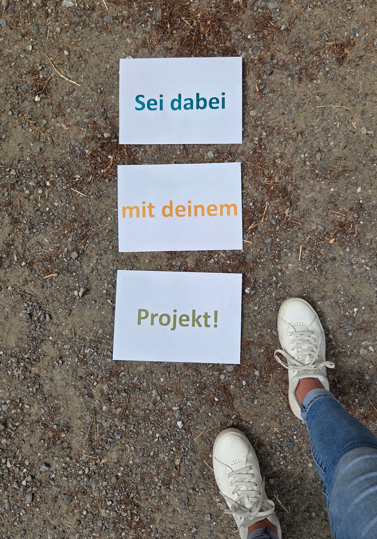 Drei weiße Karten mit deutschem Text liegen auf einer Kiesoberfläche. Die oberste Karte zeigt 'Sei dabei', die mittlere 'mit deinem' und die unterste 'Projekt!'. Rechts ist der Fuß einer Person in weißen Turnschuhen zu sehen.