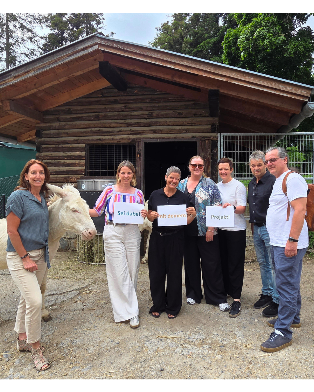 Sechs Erwachsene und ein weißes Pferd stehen vor einem Holzstall und halten Schilder mit den Aufschriften 'Seid dabei', 'mit deinem' und 'Projekt!'.