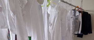 Ein Raum mit weißen Wänden hat mehrere weiße Kleidungsstücke, die an einer Stange hängen, darunter einige mit Plastikabdeckungen. Zwei Kartons voller Gegenstände stehen auf dem Boden.