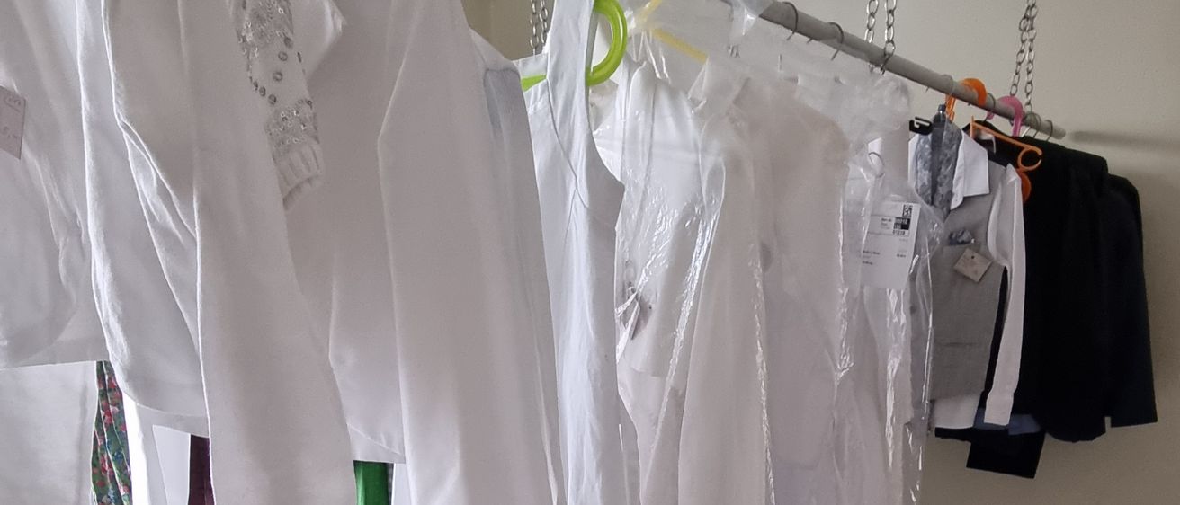 Ein Raum mit weißen Wänden hat mehrere weiße Kleidungsstücke, die an einer Stange hängen, darunter einige mit Plastikabdeckungen. Zwei Kartons voller Gegenstände stehen auf dem Boden.