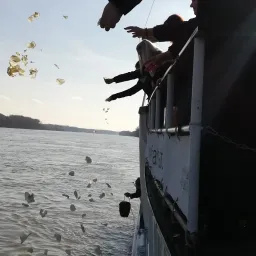 Mehrere Personen werfen auf einem Boot Gegenstände ins Wasser. Das Boot befindet sich mitten in einem großen Gewässer.