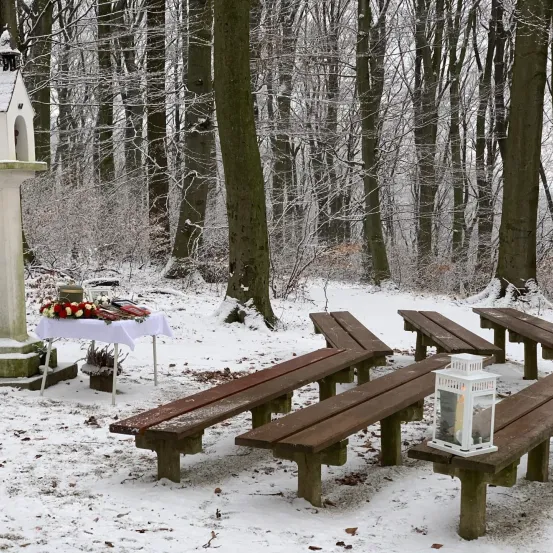 Eine Winterszene mit schneebedecktem Boden, einem kleinen Denkmal mit Blumen und Holzbänken. Eine Laterne ist auf der Bank angezündet.