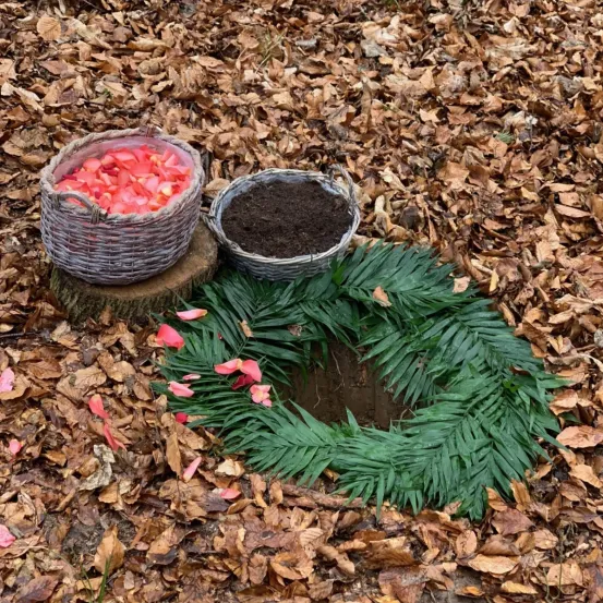 Ein Haufen roter Blütenblätter in einem Korb aus Weide, ein Korb mit Erde und ein grüner Blattkranz um ein Loch im Boden, alles auf einem Bett aus trockenen Blättern.