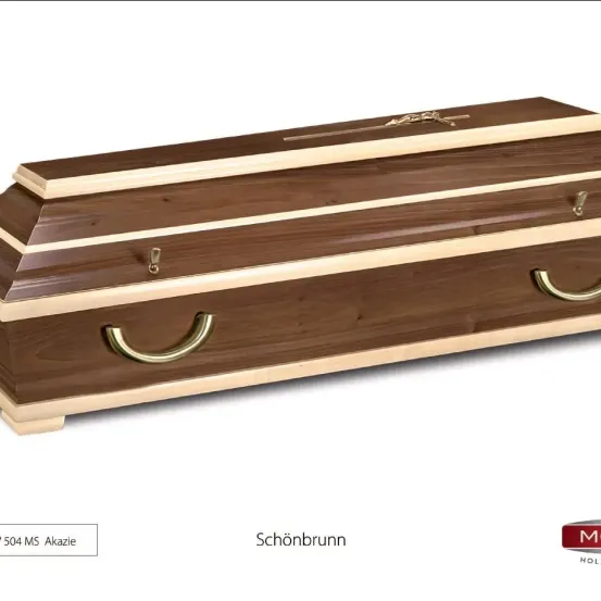 Das Bild zeigt einen hölzernen Sarg mit der Aufschrift '504 MS Akazie', goldenen Griffen und einem dekorativen Kreuz oben, vor einem weißen Hintergrund.