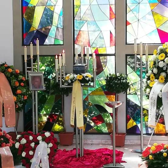 Eine Gedenkfeier mit Kerzen, Blumen und bunten Buntglasfenstern. Kerzen stehen vor den Fenstern, mit rotem Stoff auf dem Boden und verschiedenen Blumen drumherum.