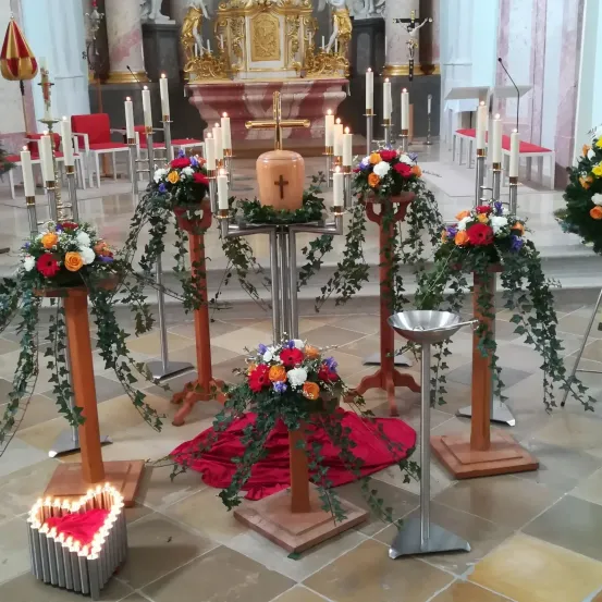 Ein Kircheninneres geschmückt mit Blumenarrangements und brennenden Kerzen. Ein großes Kreuz steht zentral, umgeben von Kerzen auf Ständern. Bunte Blumen fließen über die Ständer hinab.