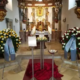 Eine Kircheninnenansicht mit Altar, Kerzen und Blumenarrangements. Es gibt zwei Blumensträuße auf beiden Seiten des Altars, in Blau gehüllt. Ein roter Stoff bedeckt den Boden.