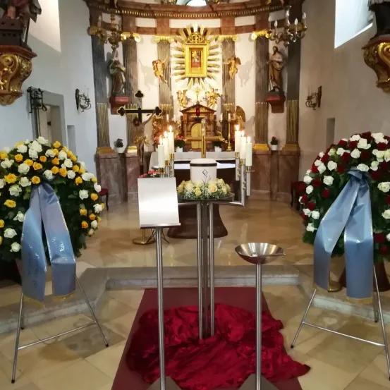 Eine Kircheninnenansicht mit Altar, Kerzen und Blumenarrangements. Es gibt zwei Blumensträuße auf beiden Seiten des Altars, in Blau gehüllt. Ein roter Stoff bedeckt den Boden.