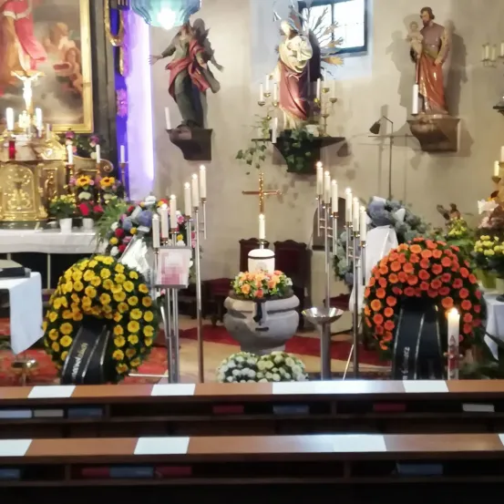 Eine Kircheninnenansicht mit Altar, Blumenarrangements, Kerzen und Statuen. Ein Kreuz und Blumen schmücken den Altar, und Statuen säumen die Wände.