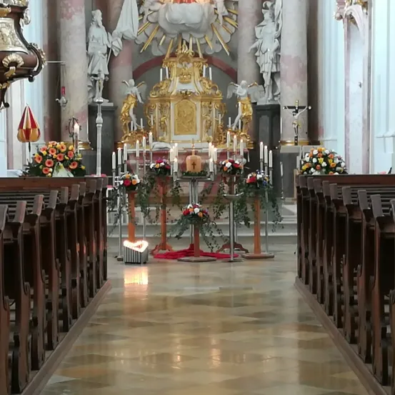 Ein Kircheninneres mit einem goldenen Altar, geschmückt mit Kerzen und Blumen, flankiert von Statuen. Reihen von Kirchenbänken sind zum Altar ausgerichtet, und dekorative Elemente schmücken die Wände.