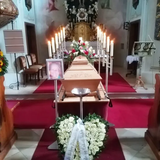 Ein Sarg mit Blumen und Kerzen in einer Kirche, gerahmtes Bild in der Nähe, rote Teppiche und Stühle im Hintergrund.