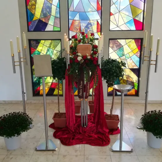 Ein Kirchenaltar-Setup mit mehrfarbigen Buntglasfenstern, mit einer zentralen Holzbox mit Blumen und Kerzen auf beiden Seiten. Bepflanzte Töpfe und Metallkerzenständer sind symmetrisch um den Altar herum angeordnet.