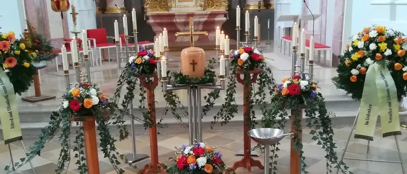 Ein dekorierter Kirchenaltar mit mehreren Kerzenständern, Blumen und einer silbernen Schüssel in der Mitte. Der Altar hat ein Kreuz und einen roten Teppich auf dem Boden.