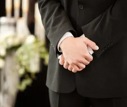 Ein Mann in einem schwarzen Anzug steht bei einer Beerdigung und hält seine Hände vor sich zusammen. Dahinter ist ein verschwommenes Bild von einer Kerze und Blumen zu sehen.