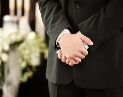 Ein Mann in einem schwarzen Anzug steht bei einer Beerdigung und hält seine Hände vor sich zusammen. Dahinter ist ein verschwommenes Bild von einer Kerze und Blumen zu sehen.