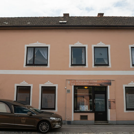 Ein beigefarbenes Gebäude mit mehreren Fenstern und einer Tür. Ein braunes Auto ist davor geparkt.