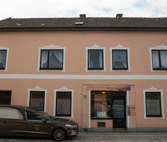 Ein beigefarbenes Gebäude mit mehreren Fenstern und einer Tür. Ein braunes Auto ist davor geparkt.