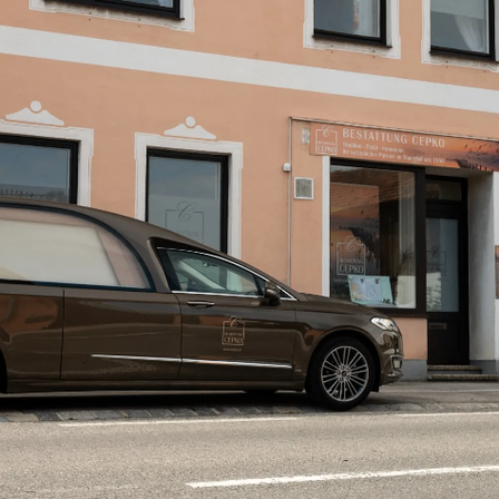 Ein brauner Leichenwagen steht vor einem Gebäude mit einem Schild, das 'Bestattung Cepko' steht. Das Gebäude hat mehrere Fenster und eine Tür. Der Leichenwagen hat ein Logo an seiner Seite.