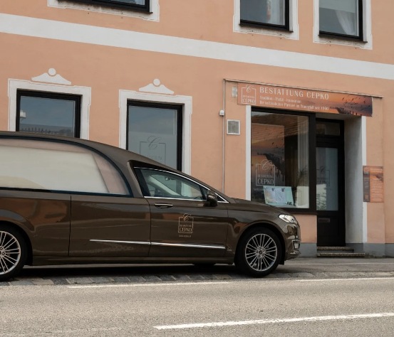 Ein brauner Leichenwagen steht vor einem Gebäude mit einem Schild, das 'Bestattung Cepko' steht. Das Gebäude hat mehrere Fenster und eine Tür. Der Leichenwagen hat ein Logo an seiner Seite.