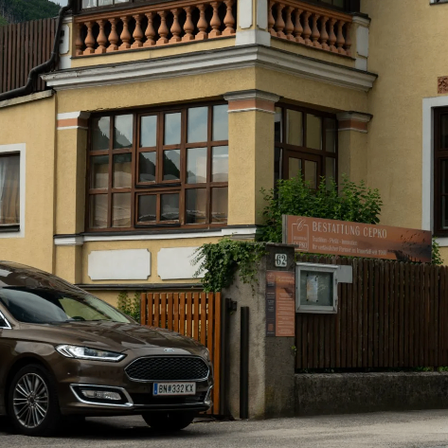 Ein braunes Auto ist vor einem gelben Gebäude mit braunen Fenstern und einem Balkon geparkt. Ein Holzzaun umgibt den Eingang, und ein Schild steht 'BESTATTUNG CEPKO'.