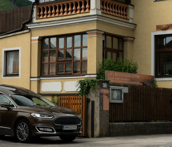 Ein braunes Auto ist vor einem gelben Gebäude mit braunen Fenstern und einem Balkon geparkt. Ein Holzzaun umgibt den Eingang, und ein Schild steht 'BESTATTUNG CEPKO'.