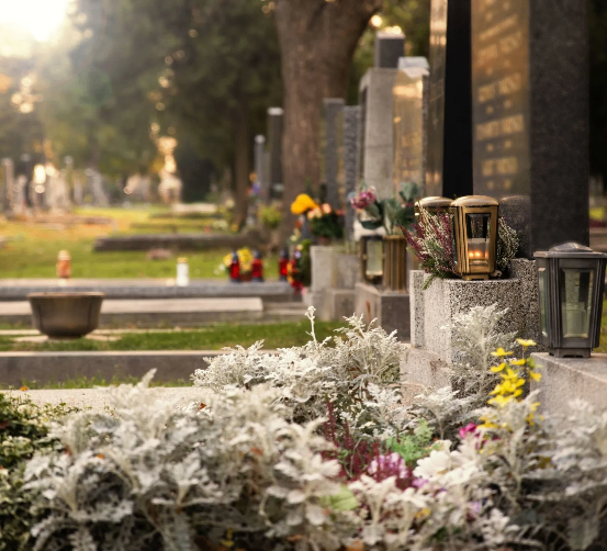 Ein Friedhof mit vielen Grabsteinen, Blumen und brennenden Laternen. Grüner Rasen und Bäume umgeben das Gelände.