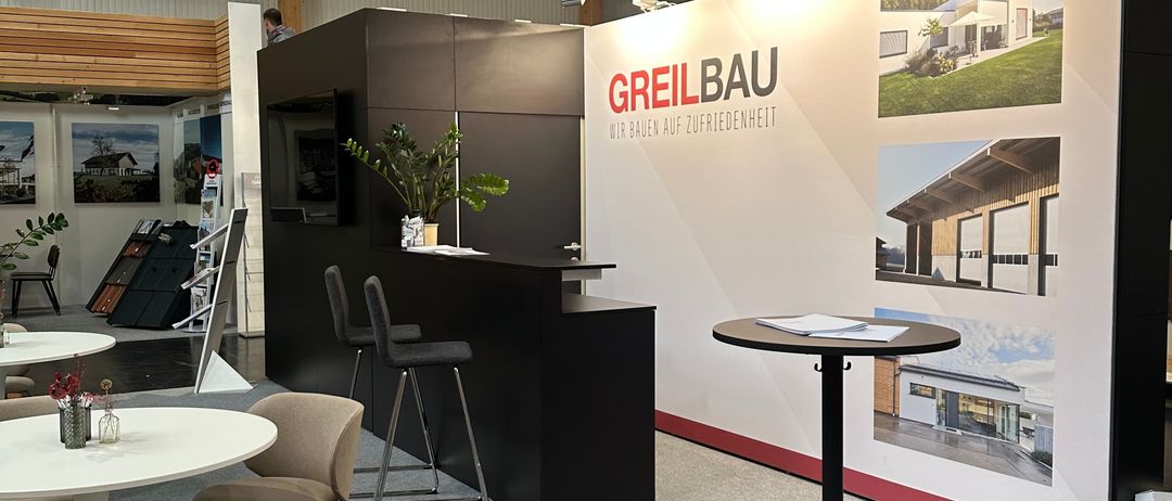 Ein Innenraum mit einem großen Banner, auf dem 'GREILBAU' steht, zeigt verschiedene Architekturdesigns. Ein runder Tisch mit Papieren steht vor dem Banner. Mehrere Tische und Stühle sind im Raum aufgestellt.