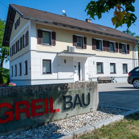 Ein weißes Gebäude mit braunem Dach, mehreren Fenstern und einem Betonschild, auf dem 'GREIL BAU' steht. Das Gebäude ist von Gras und Kies umgeben, und ein Auto ist in der Nähe geparkt.