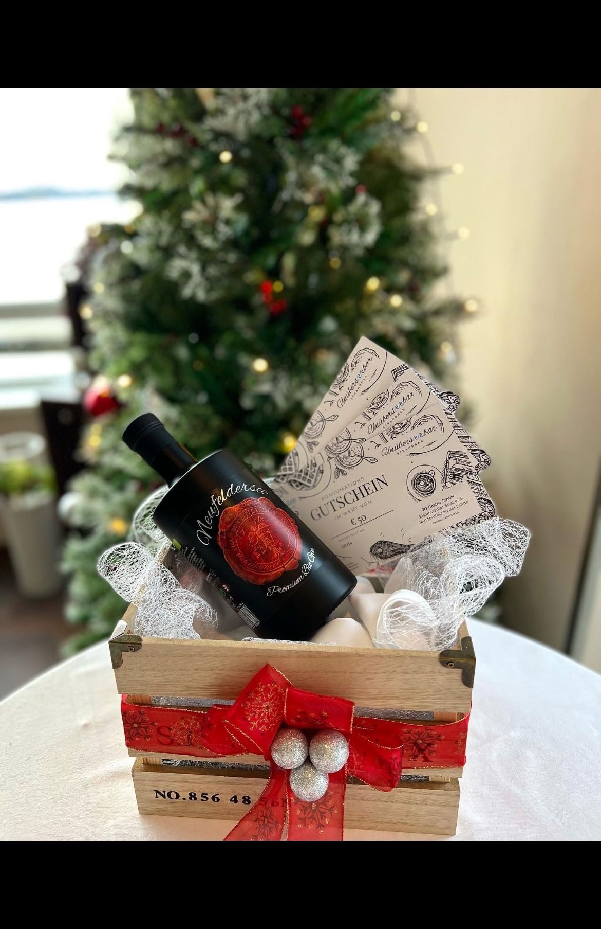 Eine Holzkiste mit einer Flasche Wein, verpackt in weißes Band und mit einem roten Band verschnürt. Die Flasche hat ein rotes Siegel darauf. Die Kiste ist mit einem Weihnachtsthema dekoriert.