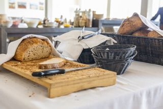 Ein Holzschneidebrett mit Brot und einem Messer steht auf einem weißen Tisch. Ein Korb mit Brot und einer Serviette steht neben dem Brett. Ein weiterer Korb steht rechts. Einige Flaschen sind auf dem Regal im Hintergrund angeordnet.